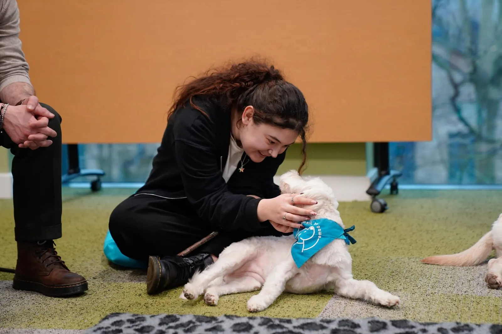 A smiling student sits on the floor cuddling a white therapy dog wearing a Moment to Paws bandana